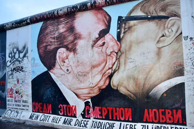 A famous colorful graffiti mural on a textured concrete wall section of the Berlin Wall. The artwork features a close-up, profile depiction of two older, bespectacled men in suits—Soviet leader Leonid Brezhnev and East German leader Erich Honecker—engaged in a passionate fraternal kiss. The background of the mural is covered in graffiti tags, and bold Russian and German text runs along the bottom border.