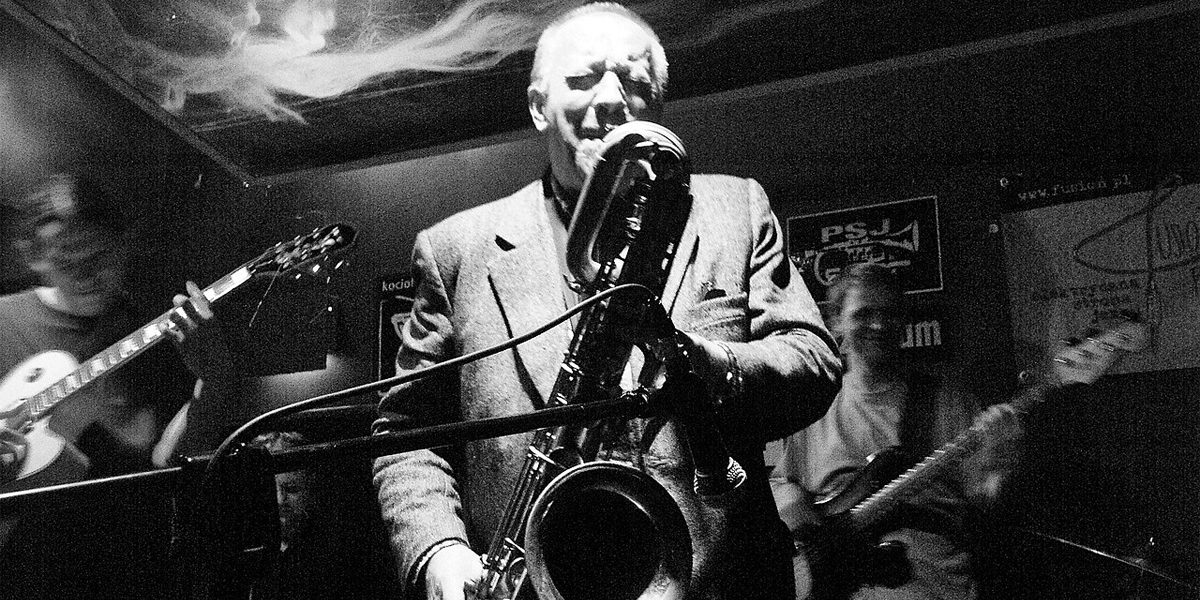 A high-contrast black-and-white photograph capturing Polish jazz musician Waldemar Kurpiński performing live on a dark, dimly lit stage. Centered in the frame, he wears a light-colored suit jacket over a dark shirt, holding a large baritone saxophone angled downward toward the microphone. His eyes are closed, showing deep concentration as he plays. Behind him, members of his jazz band are partially visible in the shadows, including a guitarist on the left and a bass player on the right.