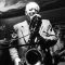 A high-contrast black-and-white photograph capturing Polish jazz musician Waldemar Kurpiński performing live on a dark, dimly lit stage. Centered in the frame, he wears a light-colored suit jacket over a dark shirt, holding a large baritone saxophone angled downward toward the microphone. His eyes are closed, showing deep concentration as he plays. Behind him, members of his jazz band are partially visible in the shadows, including a guitarist on the left and a bass player on the right.