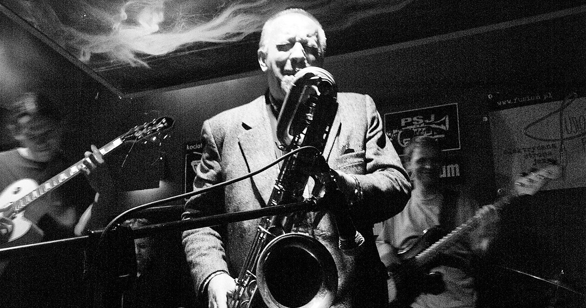 A high-contrast black-and-white photograph capturing Polish jazz musician Waldemar Kurpiński performing live on a dark, dimly lit stage. Centered in the frame, he wears a light-colored suit jacket over a dark shirt, holding a large baritone saxophone angled downward toward the microphone. His eyes are closed, showing deep concentration as he plays. Behind him, members of his jazz band are partially visible in the shadows, including a guitarist on the left and a bass player on the right.